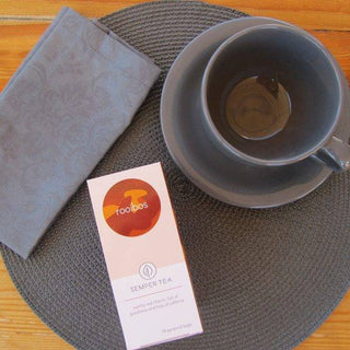 Semper Tea Rooibos en caja sobre mantel gris con taza vacía.